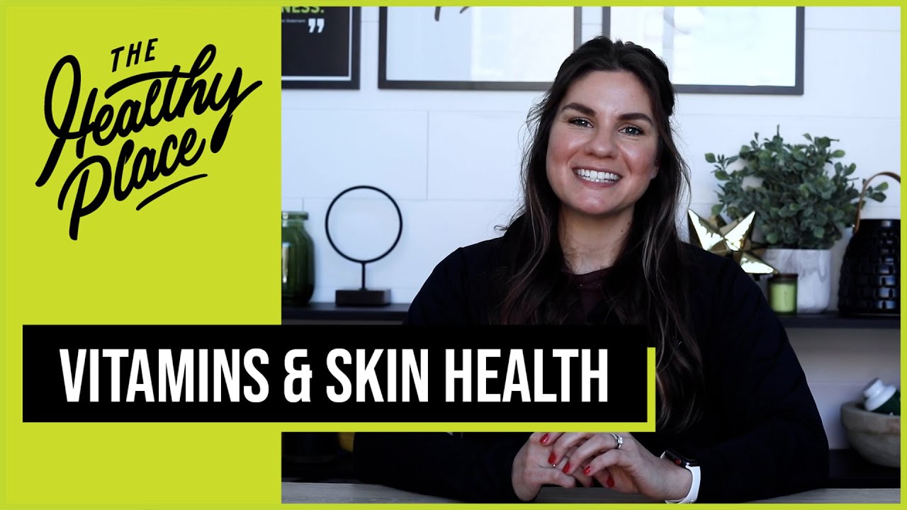 A smiling woman sits at a table with a modern background. A green overlay says "The Healthy Place," and a black banner reads "Vitamins & Skin Health."