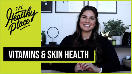 A smiling woman sits at a table with a modern background. A green overlay says "The Healthy Place," and a black banner reads "Vitamins & Skin Health."