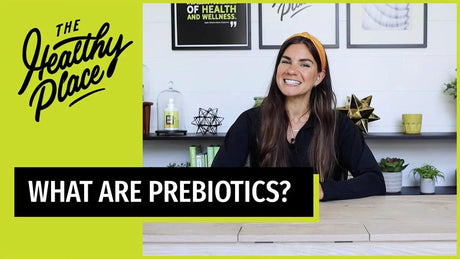 A woman in a 'Find Your Healthy Place' zip-up and orange headband sits at a table with a wellness-themed background. Text overlay reads 'WHAT ARE PREBIOTICS?'