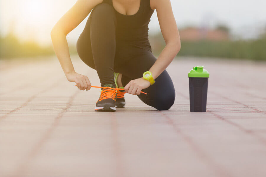 A woman dressed in black athletic wear kneels on a paved outdoor path, tying the bright orange laces of her running shoes. She wears a neon green sports watch, and a black and green protein shaker bottle sits beside her. 