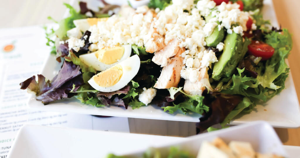 A fresh salad served at Fit Fresh Café in Madison, WI, featuring leafy greens, hard-boiled egg, feta cheese, cherry tomatoes, and avocado, with a menu in the background.