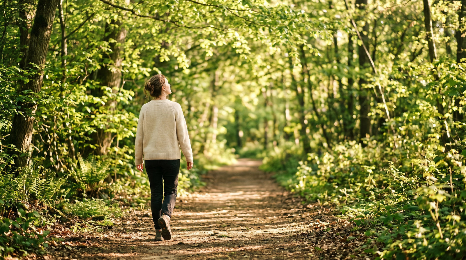 A person walks down a sun-dappled wooded path on a clear spring morning, head tilted up toward the tree canopy, breathing in fresh air. Soft natural light filters through green leaves overhead.