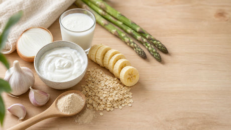 A spread of prebiotic and probiotic foods including yogurt, kefir, oats, banana, garlic, onion and asparagus on a wood surface.