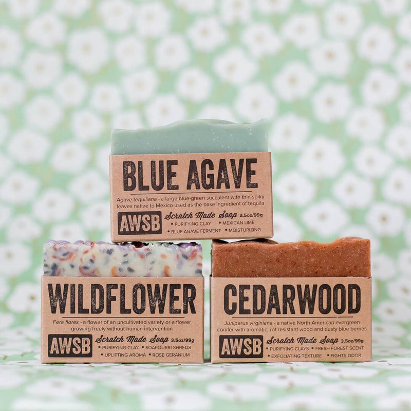 Three artisan bar soaps stacked against a floral green background, each wrapped in brown kraft paper with bold black lettering. The soaps are minty-green, pale pink, and brown in color, and they are stacked into the shape of a small pyramid. 