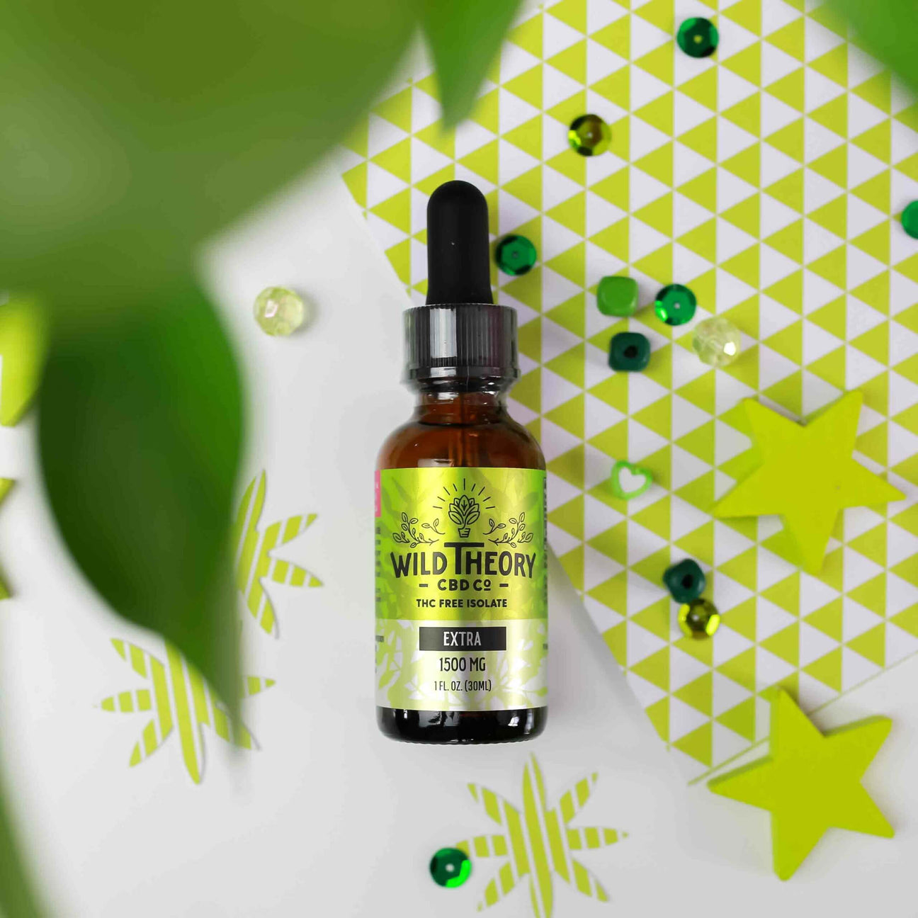 A bottle of Wild Theory CBD Isolate Extra 1500 MG is placed on a geometric green and white patterned background. Green sequins and star-shaped cutouts are scattered around, with blurred leaves in the foreground.