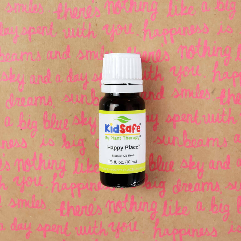 A small dark glass bottle of "KidSafe Happy Place" essential oil blend by Plant Therapy stands against a background with pink cursive writing. The label indicates it's a 10ml (1/3 fl. oz.) bottle, designed as a child-safe essential oil blend.