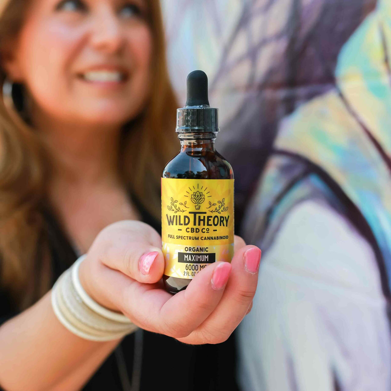 A smiling woman holds out a bottle of Wild Theory Full Spectrum CBD oil with a yellow label. The focus is on the product, while the woman and a colorful mural in the background are slightly blurred.