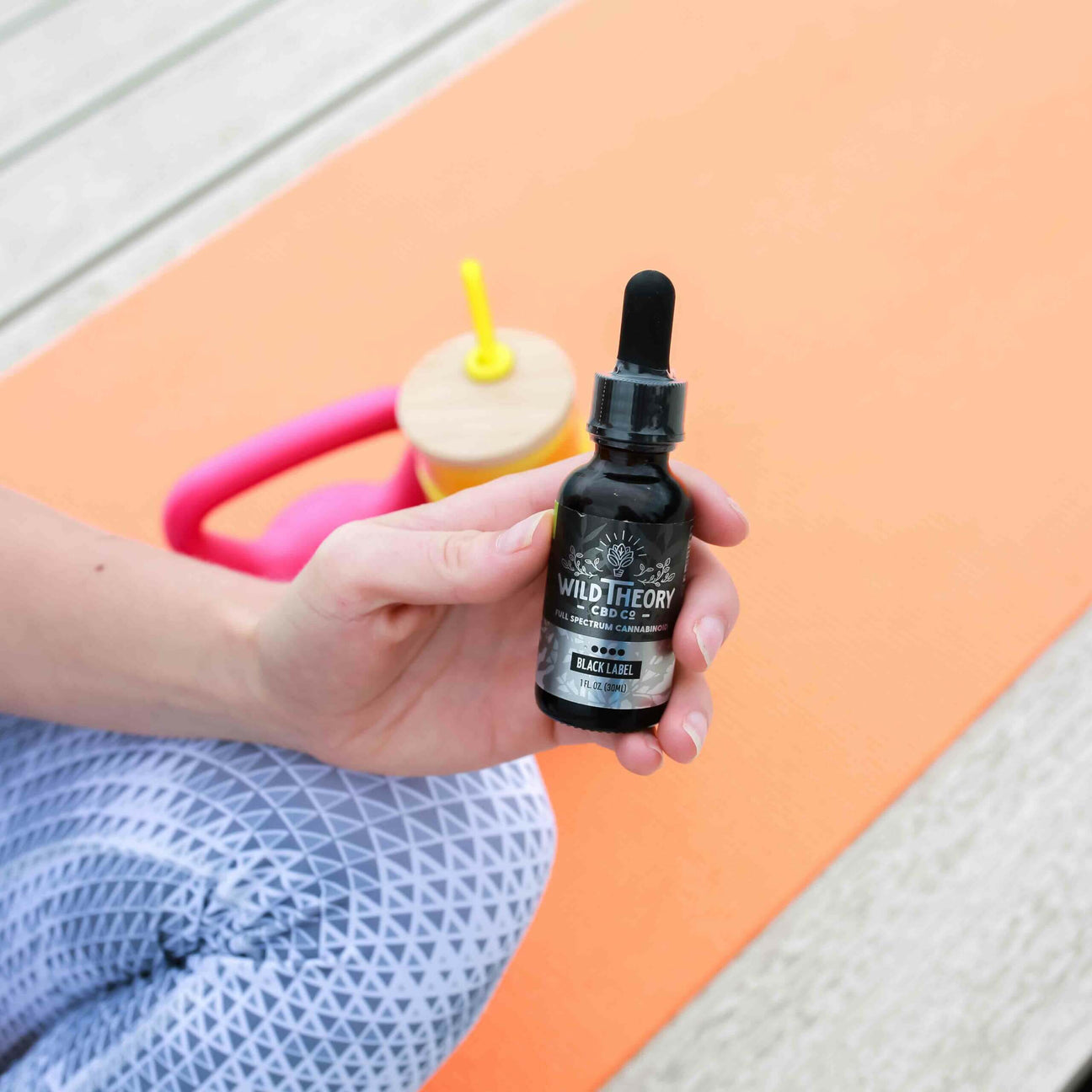 A person holds a bottle of Wild Theory CBD Co. Black Label Full Spectrum Cannabinoid Hemp Extract while sitting on an orange yoga mat. A pink and yellow water bottle is visible in the background.