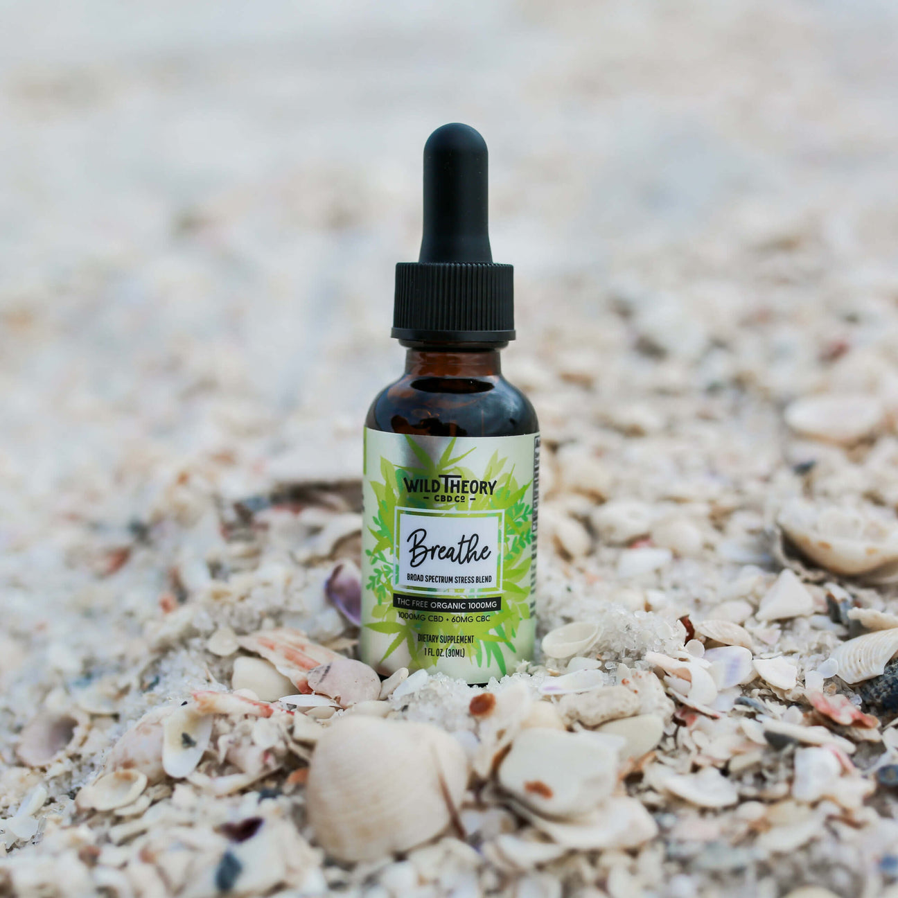 A bottle of Wild Theory CBD Co. Breathe Broad Spectrum Stress Blend sits on a bed of crushed seashells. The label highlights that it is THC-free, organic, and contains 1000mg CBD with 600mg CBG.