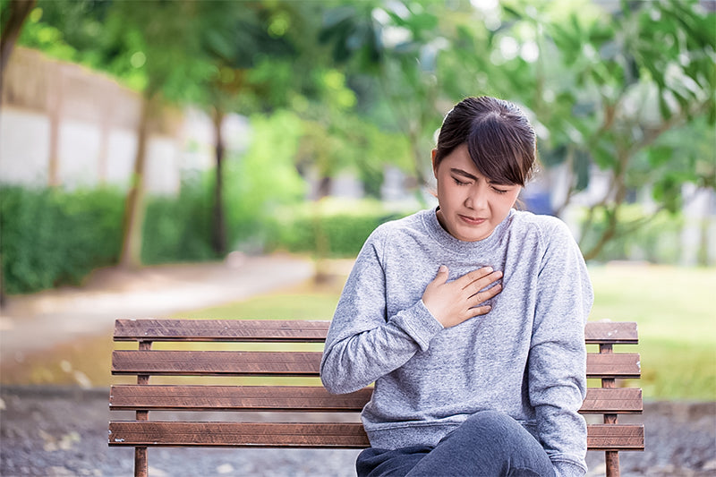 A young woman in a gray sweatshirt sits on a wooden bench in a park, pressing her hand to her chest with a pained expression. The lush background greenery contrasts with her discomfort, suggesting she may be experiencing heartburn or acid reflux.