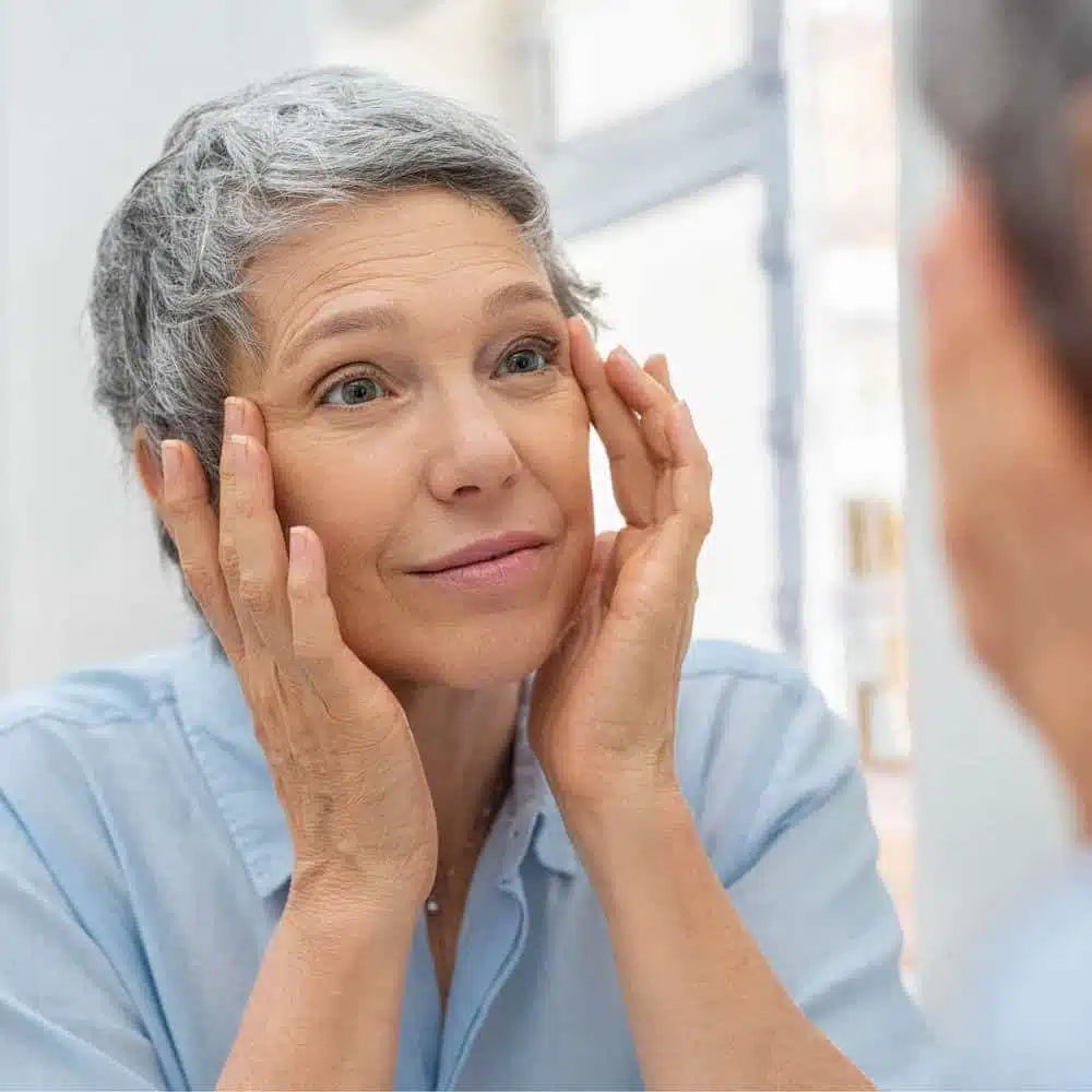 A mature woman with short gray hair gently touches her face while looking into a mirror. She wears a light blue button-up shirt and has a calm, content expression. The bright, natural lighting highlights her smooth skin.