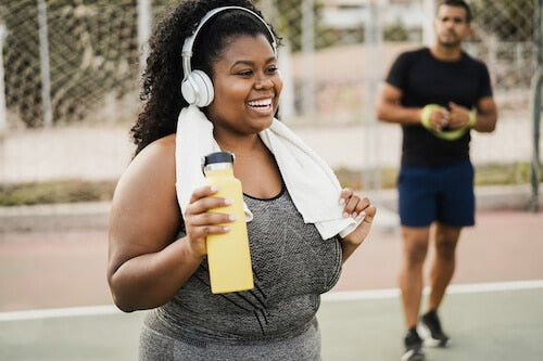 Smiling woman with curly hair, wearing headphones and a workout outfit, holds a yellow water bottle and has a white towel draped around her neck. She appears refreshed after exercising on an outdoor basketball court.