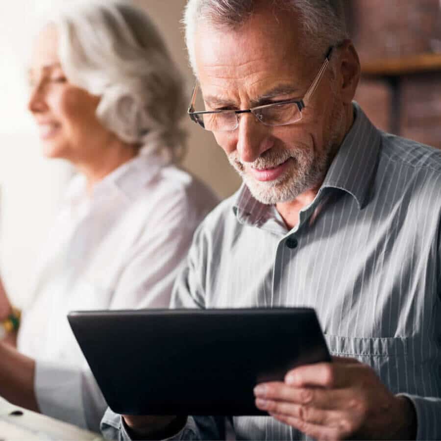 An aged man with a white beard and glasses smiles, looking at a tablet. He wears a striped button-down shirt. In the background, a woman with short white hair in a white blouse is slightly out of focus, engaged in an activity in a bright, modern setting.