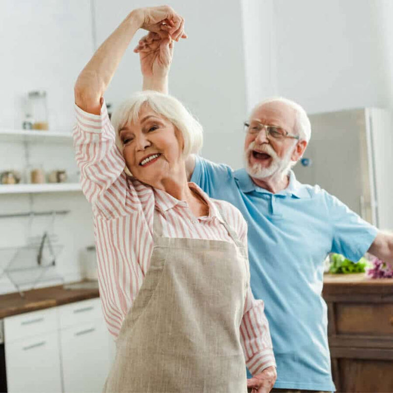 A smiling elderly couple dances in a bright kitchen. The woman, wearing a beige apron, lifts her arm as the man in a light blue polo twirls her. Shelves with jars and kitchen items are in the background, creating a warm, cheerful atmosphere.