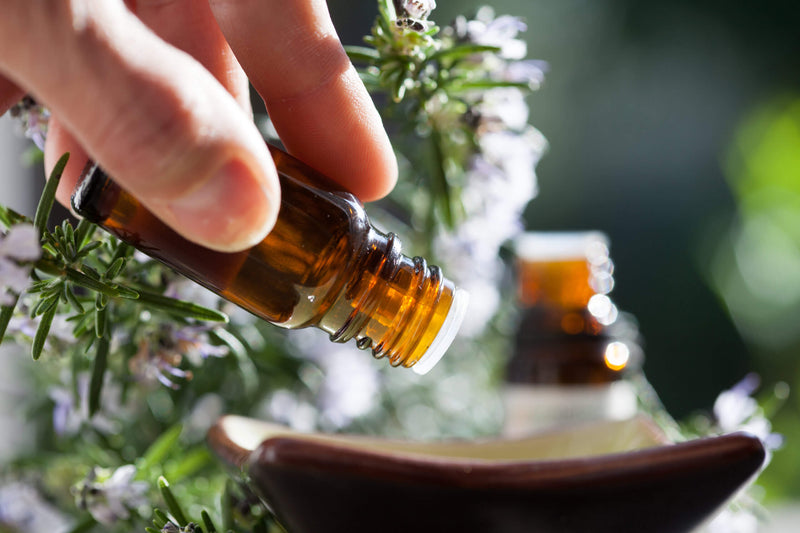 A hand carefully pours essential oil from a small amber glass bottle into a dish, with a blurred background of fresh rosemary and blooming flowers. The scene is bathed in natural sunlight, emphasizing the purity and natural essence of the oil.