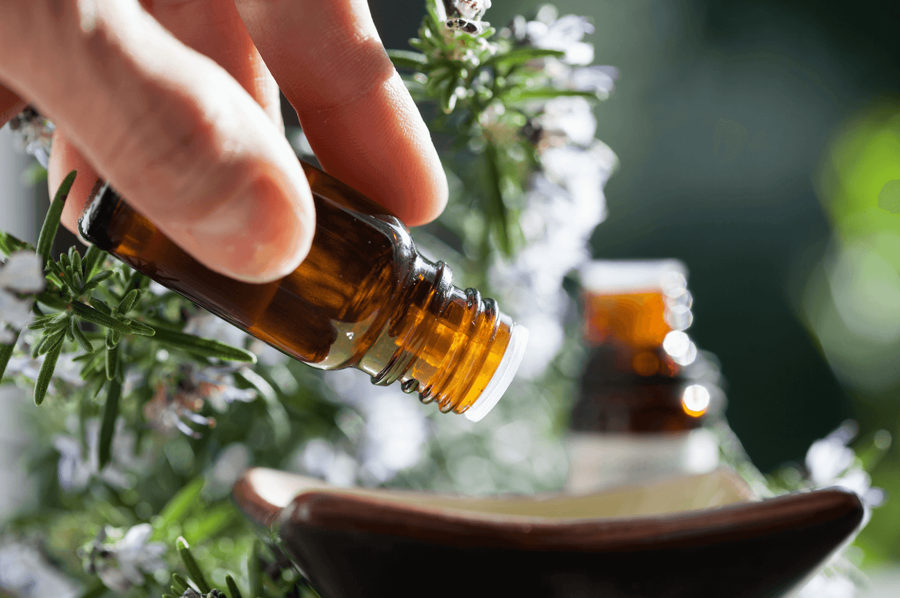 A hand carefully pours essential oil from a small amber glass bottle into a dish, with a blurred background of fresh rosemary and blooming flowers. The scene is bathed in natural sunlight, emphasizing the purity and natural essence of the oil.