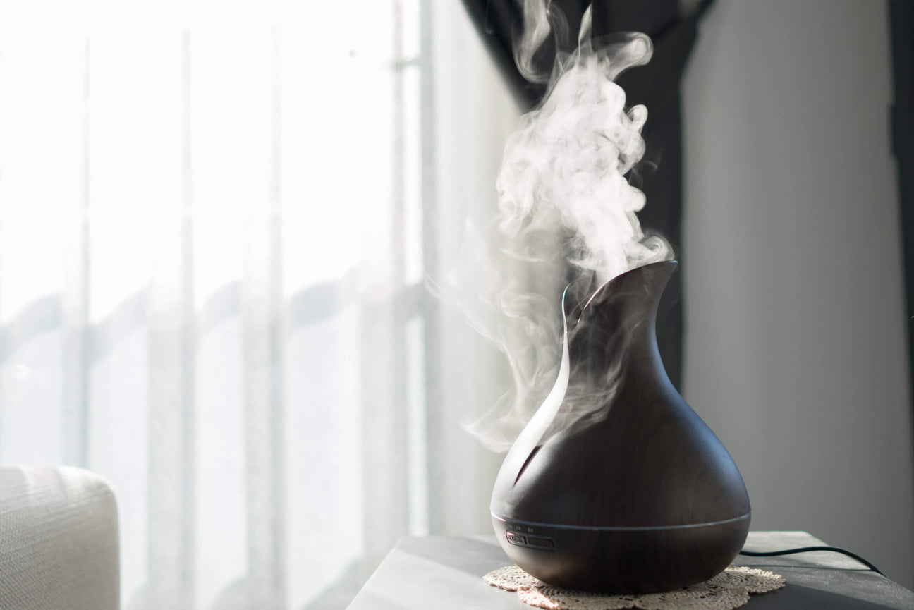 A sleek, dark wood-grain essential oil diffuser releasing a steady stream of aromatic mist into a softly lit room. Positioned on a small table near a window with sheer curtains, the diffuser adds a calming ambiance to the space.