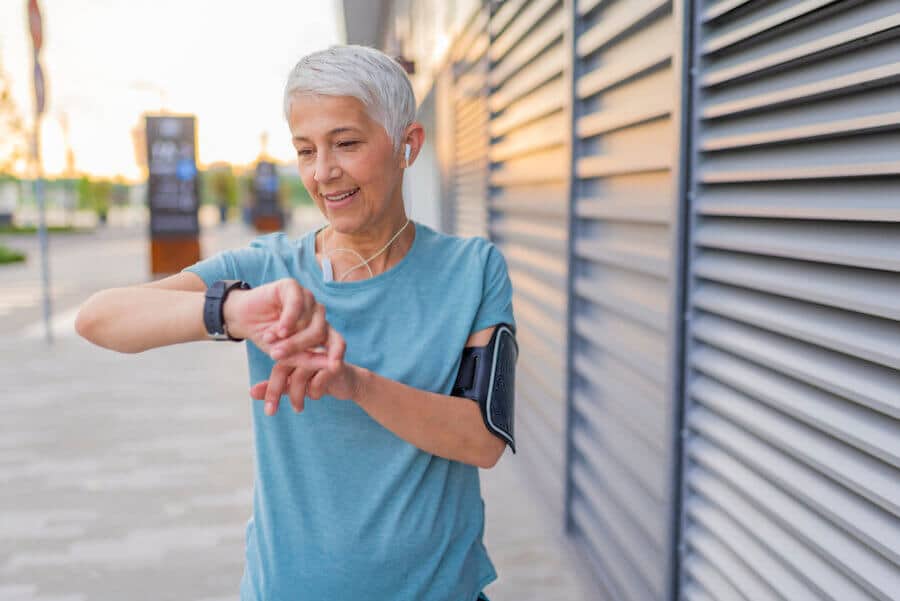 A smiling older woman with short gray hair checks her smartwatch while on a walk. She wears a light blue athletic shirt, earbuds, and an armband for her phone. The background features a modern outdoor setting with soft evening sunlight.