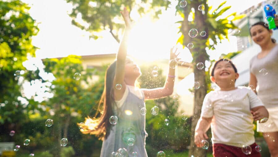 Two joyful children play outside in the sunlight, reaching for floating bubbles. A woman, possibly their mother, watches nearby with a smile. The lush green background and warm light create a cheerful, carefree atmosphere.