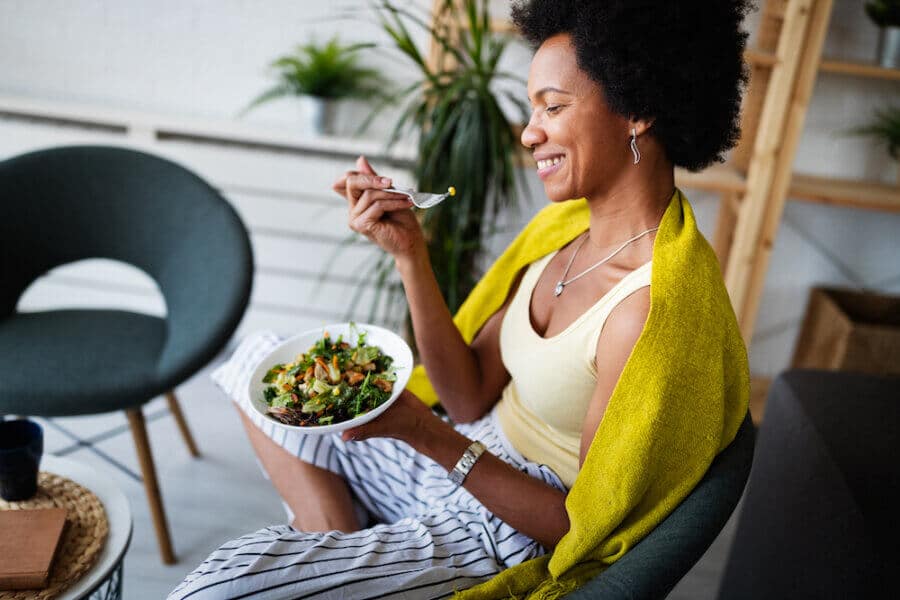 A smiling woman with curly hair enjoys a fresh salad while sitting in a cozy space. She wears a yellow tank top with a green shawl draped over her shoulders. The setting includes plants and comfortable furniture, creating a relaxed atmosphere.