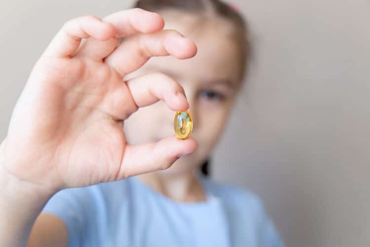 A young child in a light blue shirt holds up a translucent yellow omega-3 supplement capsule between their fingers. The child's face is blurred in the background, focusing attention on the softgel.