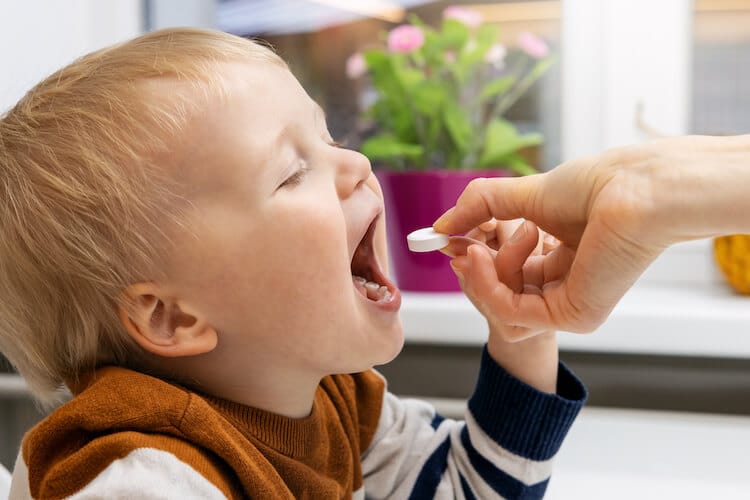 A young child with short blond hair and a striped sweater opens their mouth wide as an adult hand offers a white chewable tablet. The background features a softly lit indoor setting with a potted plant and flowers on a windowsill.