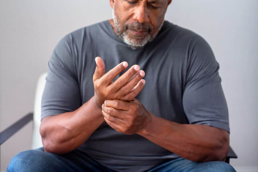      middle-aged man with a gray beard and muscular build sits in a chair, wearing a dark gray t-shirt. He looks down at his hand with discomfort, massaging it with his other hand, suggesting pain or stiffness.