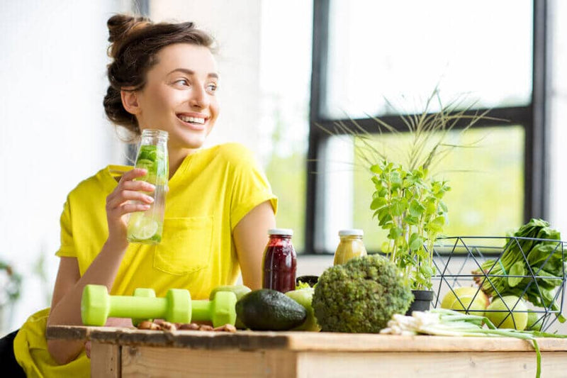 A smiling woman in a bright yellow shirt holds a glass of infused water while sitting at a table filled with fresh vegetables, herbs, and juices. Green dumbbells and a vibrant indoor setting suggest a focus on health and wellness.