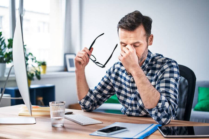 A man in a checkered shirt sits at a desk, rubbing his eyes while holding his glasses in one hand. His workspace includes a computer, a notebook, and a glass of water. His posture indicates fatigue or stress.