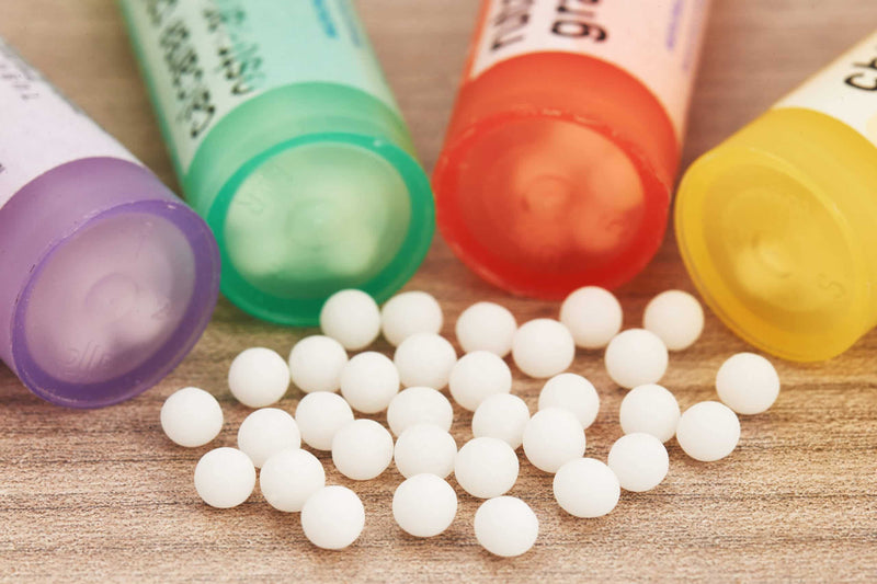 A close-up of small white homeopathic pellets scattered on a wooden surface, with colorful tubes in purple, green, red, and yellow lying nearby. The vibrant bottles contrast with the neutral background, emphasizing alternative medicine.