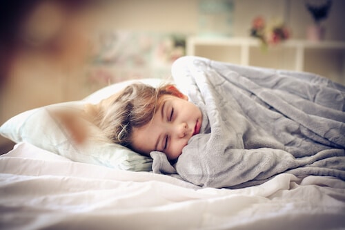 A young child sleeping soundly in bed, wrapped in a soft gray blanket. Their face looks peaceful and relaxed, with a cozy bedroom setting in the background. Natural light gently illuminates the space, creating a warm and restful atmosphere.