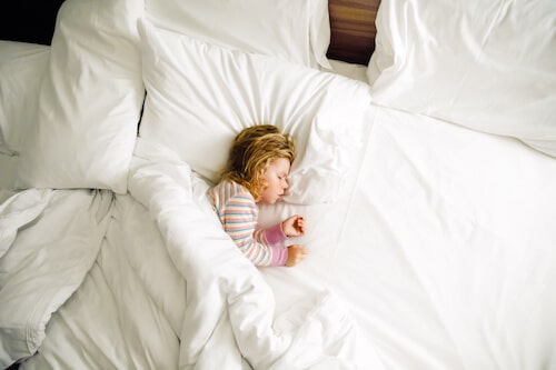 A young child sleeping peacefully in a large bed with white pillows and blankets. The child is wearing striped pajamas and is curled up comfortably under the covers. The scene has a soft, warm glow, creating a cozy and restful atmosphere.