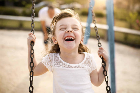 A young girl laughing joyfully while swinging on a swing set in a sunny park. She is wearing a white lace-trimmed shirt and has a carefree expression, with blurred greenery and a figure in the background.