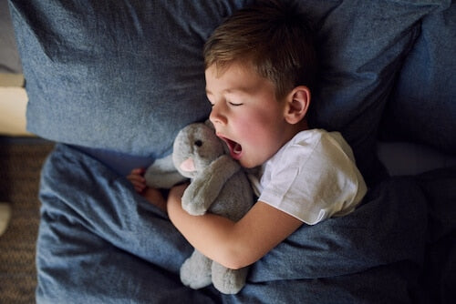 A young boy in a white t-shirt yawning while cuddling a soft gray stuffed bunny, lying in bed with deep blue bedding. The dim lighting and cozy atmosphere suggest bedtime, creating a calm and restful scene.