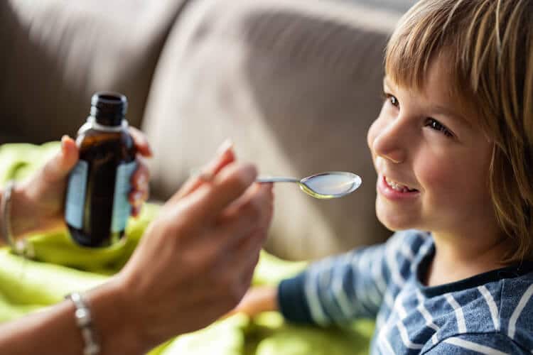 A smiling young child with shoulder-length blond hair and a striped blue sweater sits on a couch, looking at an adult's hand holding a spoonful of liquid supplement. The other hand holds a small dark glass bottle, likely containing the multivitamin.