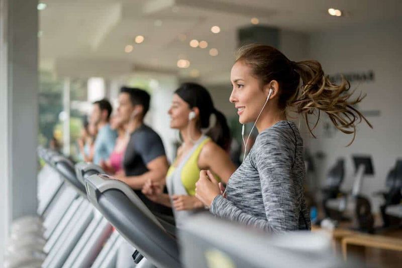 A group of people running on treadmills in a gym, focusing on a smiling woman in a gray workout top with her hair tied back. She is wearing earbuds and appears to be enjoying her workout. Other gym-goers in athletic wear are exercising in the background.