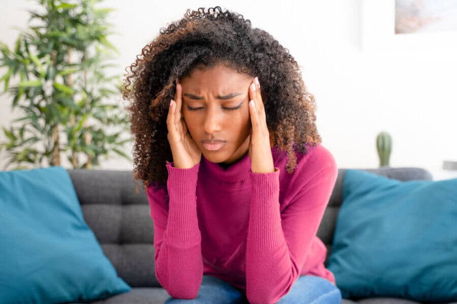 A young woman with curly hair sits on a couch, pressing her fingers to her temples with a pained expression suggesting she has a headache. She wears a burgundy sweater, and the background features a cozy living space with plants and blue pillows.