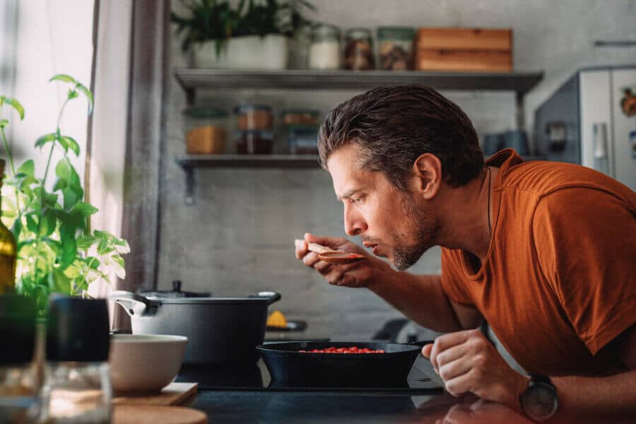 A man in an orange t-shirt leans over a stovetop, tasting food from a spoon. In front of him is a skillet full of red ingredients. The kitchen has a cozy, rustic feel with open shelves, potted plants, and natural light.