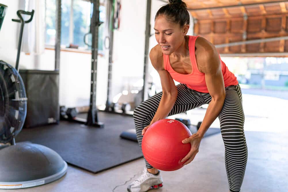  A fit woman in a bright coral tank top and striped leggings is in a home gym, holding a red medicine ball in a squat position. She appears focused and determined as she engages in a workout. 