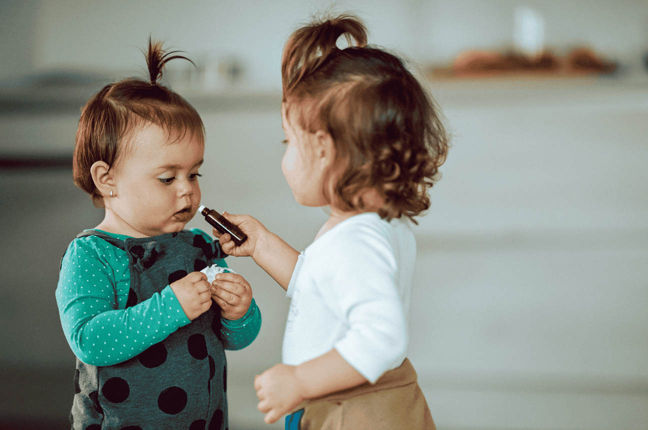 Two toddlers play together in a cozy home setting. One child holds a small essential oil bottle and offers it to the other child. The moment captures their curiosity and interaction in a warm, softly lit environment.