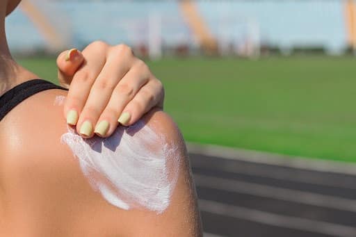 A person applying sunscreen to their shoulder with a green field and track in the background, emphasizing outdoor skin protection under sunny conditions.