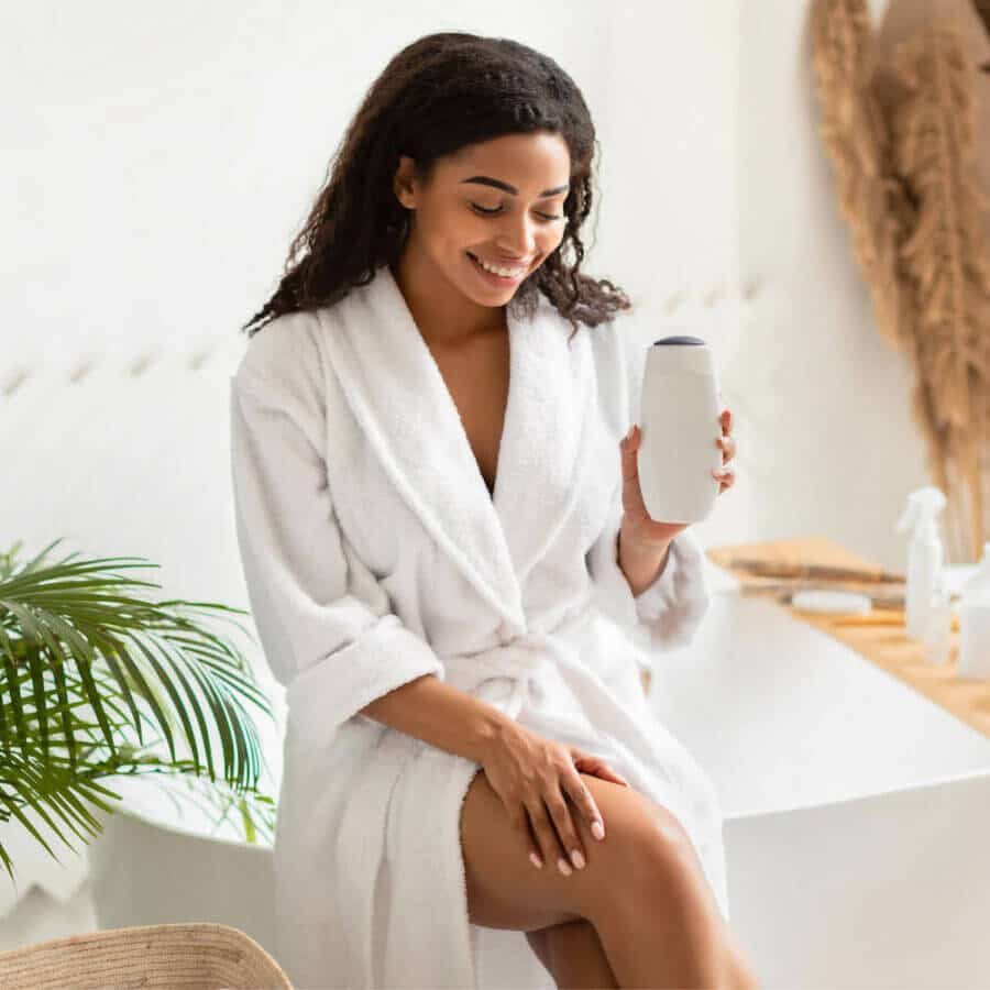 A smiling woman in a plush white robe sits near a bathtub, holding a sleek white bottle of skincare or beauty product. The bathroom has a serene, spa-like feel with natural decor, plants, and soft lighting, suggesting a self-care routine.