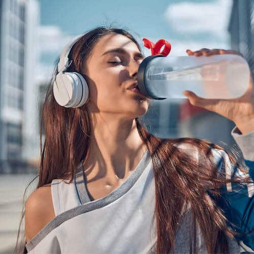 A young woman with long brown hair drinks water from a sports bottle while wearing white headphones. She is dressed in a casual off-shoulder workout top, and the background features an urban setting with modern buildings under a bright blue sky.