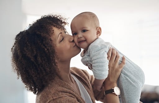 A loving mother kissing her smiling baby on the cheek while holding them up. The baby is dressed in a light gray onesie, and the setting is a softly lit indoor space, creating a warm and tender moment.