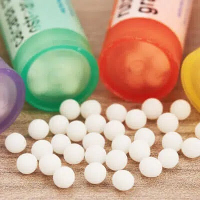 Close-up of small, round white homeopathic pellets scattered on a wooden surface, with four colorful plastic tubes — purple, green, red, and yellow — lying nearby.