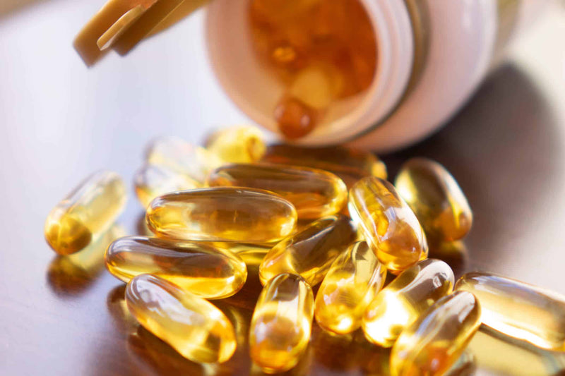 A spilled bottle of golden, translucent vitamin D softgel capsules on a reflective wooden surface. The capsules catch the light, highlighting their smooth texture and rich amber color.