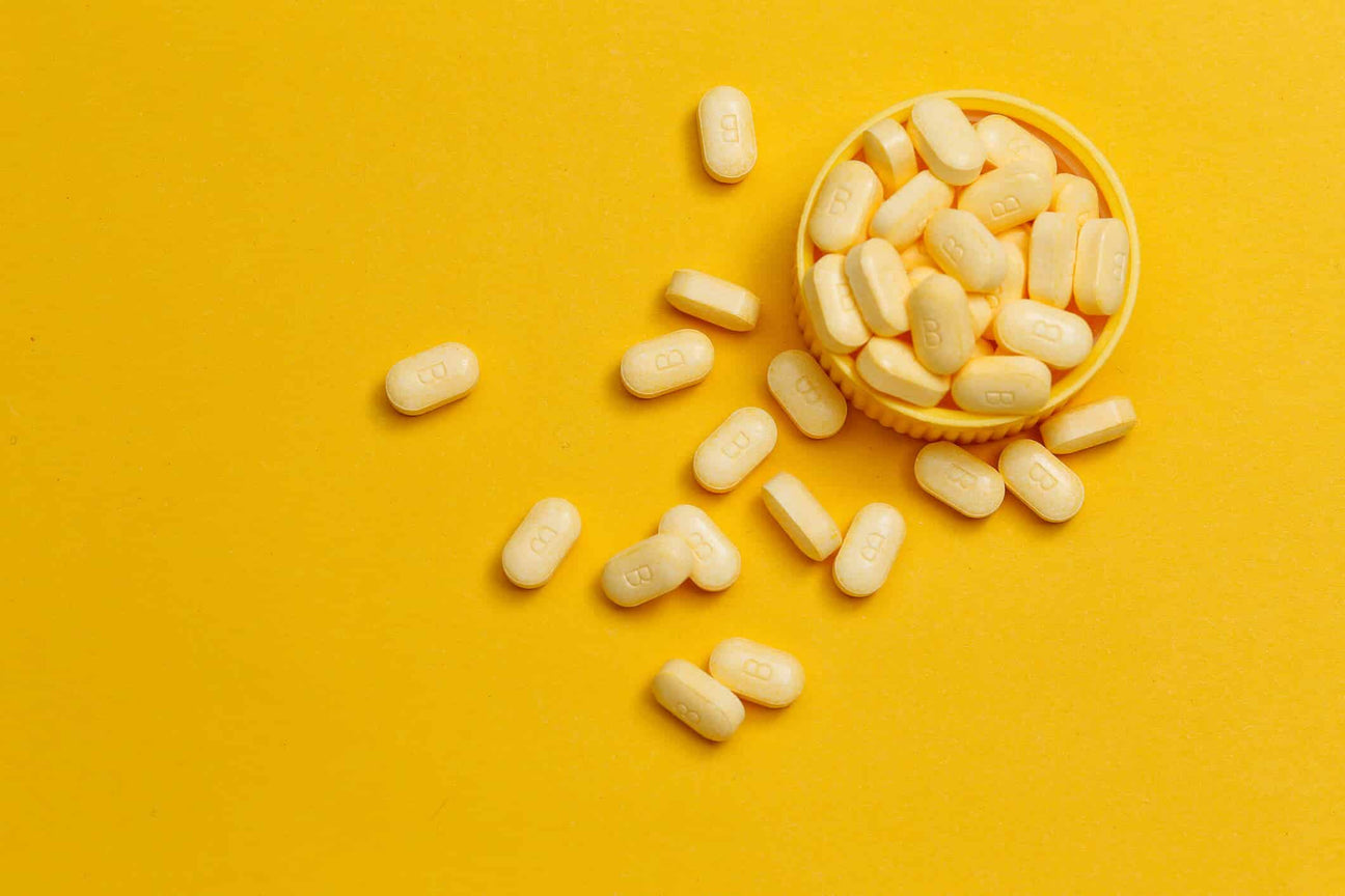 A small yellow cap filled with yellow vitamin B tablets, with several pills scattered around it on a bright yellow background. Each tablet is imprinted with the letter "B," emphasizing its nutritional content.