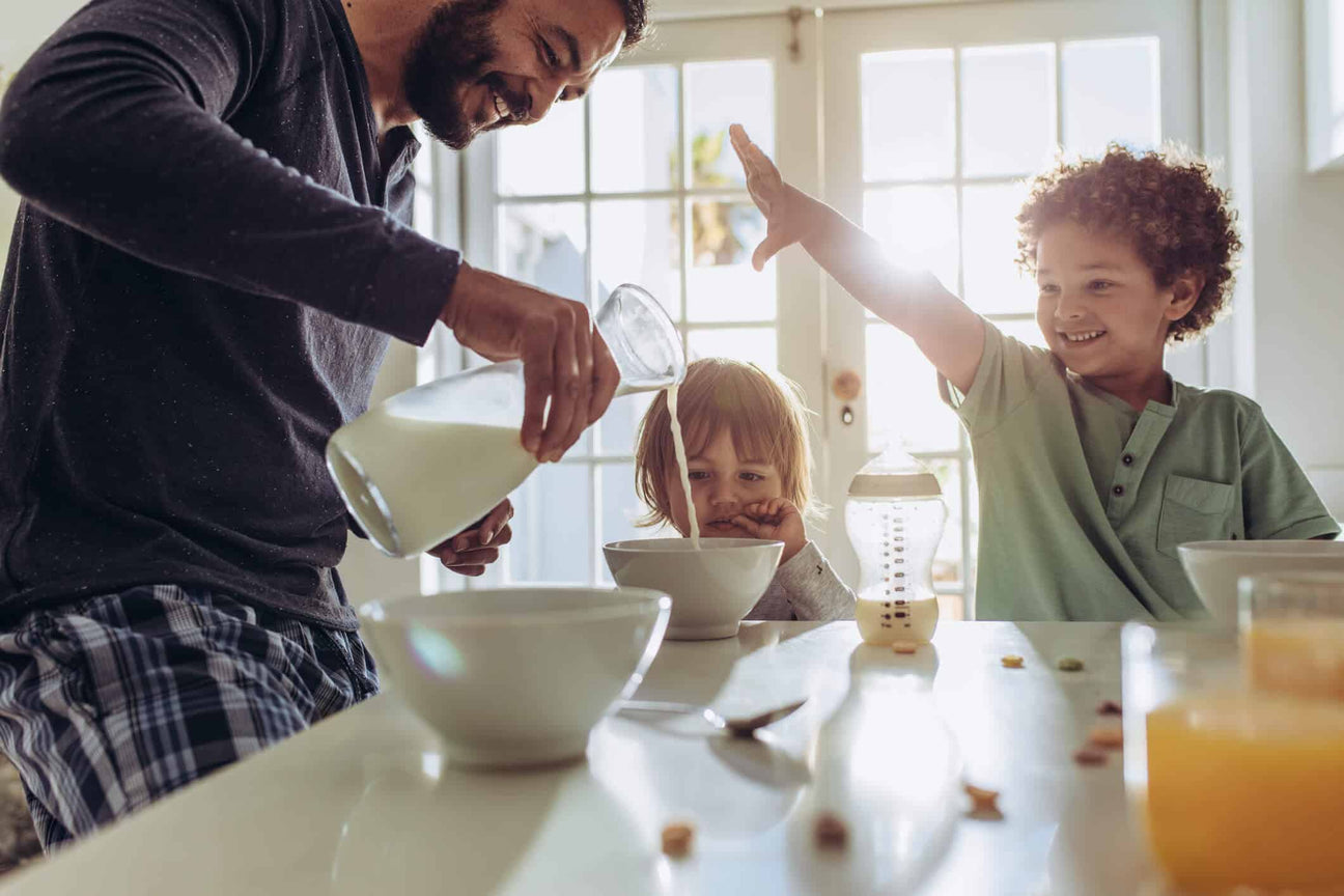 A father pours milk into a bowl for his young children at the breakfast table. One child watches curiously, while the other excitedly raises their hand. Sunlight streams through the windows, creating a warm and joyful morning scene.