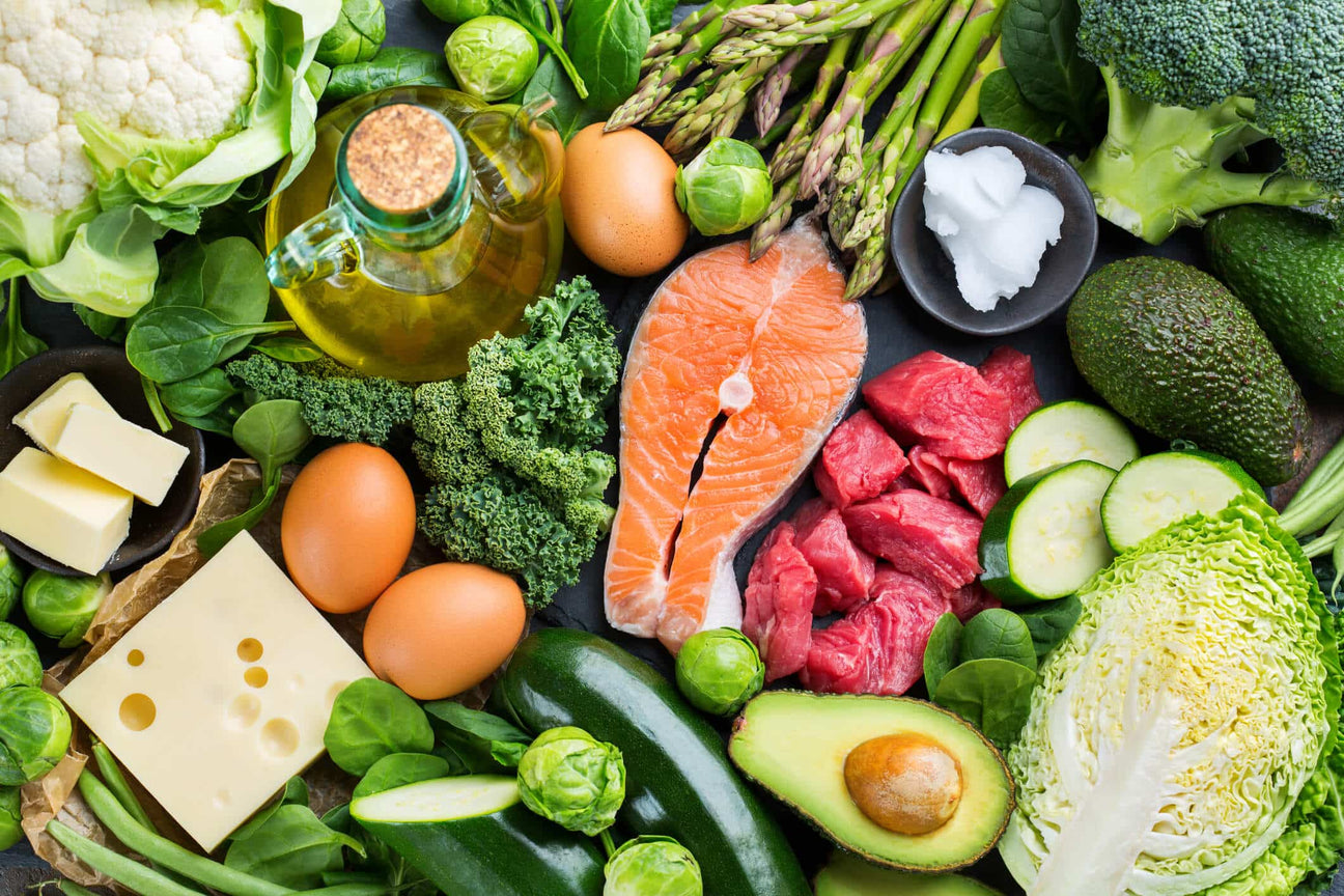 A colorful assortment of whole foods rich in omega fatty acids, including salmon, avocado, eggs, cheese, butter, olive oil, and green vegetables like kale, Brussels sprouts, and zucchini. The spread highlights nutrient-dense sources of healthy fats.