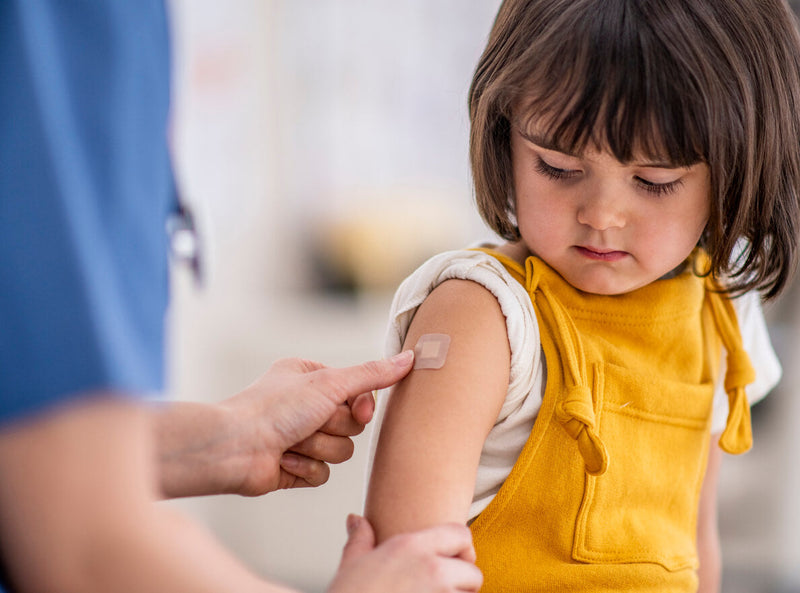 A young girl wearing mustard yellow overalls looks down at her arm as a healthcare professional in blue scrubs applies a small bandage. The child’s expression is calm but focused, showing a moment of gentle care in a medical or first-aid setting. 